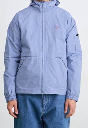 Mann trägt eine hellblaue Jacke mit Reißverschluss und Kapuze sowie einem Logo-Patch auf der Brust, kombiniert mit blauen Denim-Jeans, steht vor neutralem Hintergrund.