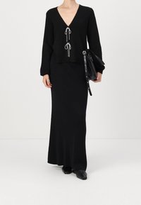 Černý pletený top s výstřihem do V a detailem se stahovací šňůrkou, v kombinaci se sladěnou dlouhou černou sukní. Modelka drží černou tašku a má na sobě černé boty.