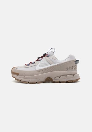 Baskets de sport beige clair et blanches avec une semelle épaisse, le logo Nike blanc, des lacets rouges et bleus et un détail texturé au talon, vues de côté.