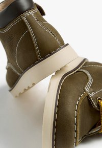Bottes de cheville en cuir marron avec des coutures blanches, une semelle en caoutchouc beige et un design courbé. Comprend une boucle de lacet pour une touche de détail supplémentaire.