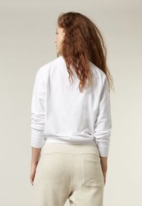 Felpa bianca in cotone con polsini e bordo a costine, abbinata a pantaloni della tuta beige chiaro con dettagli sulla tasca posteriore.