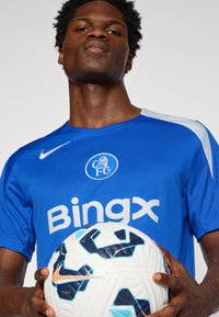 Blå fodboldtrøje med hvide accenter, med "Bingx" logo og Chelsea FC’s skjold. Spilleren holder en hvid og blå Nike fodbold.