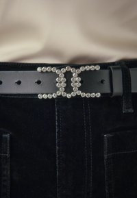 Black leather belt featuring a decorative buckle adorned with clear crystals, shaped in an interlocking 'C' design. Textured black fabric visible below.