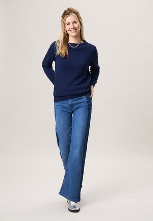 Blonde vrouw die glimlacht, draagt een marineblauw trui, blauwe wijde jeans, zilveren laarzen en een ketting, staand tegen een effen lichte achtergrond.