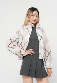 Fleece zip-jakke med et rosa og hvitt diamantmønster, svarte detaljer, og høy krage, kombinert med en grå topp og plissert skjørt.