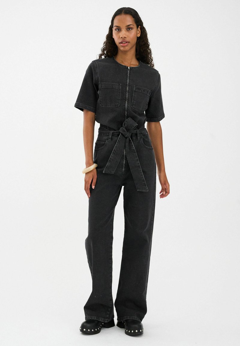 Čierny denimový overal s krátkymi rukávmi, zapínaním na zips, dvoma náprsnými vreckami a opaskom v páse. Rozšírené nohavice a hladká textúra so subtilnými detalmi prešitia.