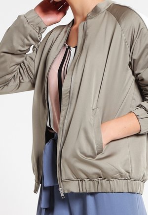 Henkilö pukeutunut beigeen vetoketjulliseen takkiin raidallisen pinkin, mustan ja valkoisen päällä, käsi takin taskussa.