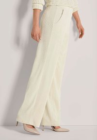 Persoon in crème kleurige wijde ribfluwelen broek, crème gebreide trui en beige spitse hoge hakken staat tegen een lichte muur.