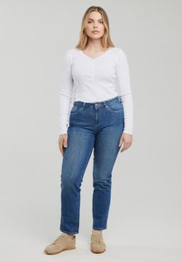 Kobieta stojąca zwrócona przodem, ubrana w białą koszulę z długim rękawem, niebieskie dżinsy i beżowe trampki na jednolitym jasnoszarym tle.