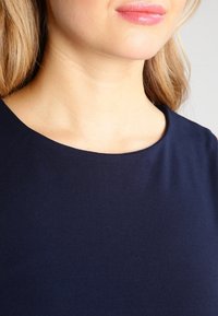 Blond kvinna som bär en marinblå topp med rund nacke, som visar hennes nedre ansikte, hals och övre bröst.