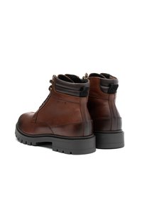 Botas de tobillo de cuero marrón con una textura suave, punteras redondas, diseño de cordones y una gruesa suela de goma negra. Presentan ojales de metal y detalles de costura.