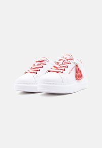 Λευκά αθλητικά παπούτσια με κόκκινα κορδόνια, λογότυπα Karl Lagerfeld στα πλάγια και στη γλώσσα, και ένα κόκκινο γαντζωτό με σχήμα καρδιάς προσαρτημένο σε ένα από τα παπούτσια.