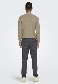 Beiger Strickpullover für Herren mit hohem Kragen, kombiniert mit dunkelgrauen karierten Hosen mit zwei Gesäßtaschen. Abgerundet mit hellen Schuhen.