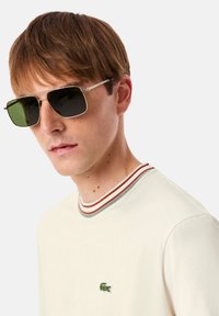 Lunettes de soleil en métal avec des verres teintés verts, de forme rectangulaire, et des accents métalliques. Portées avec une chemise crème dotée d'un col rayé et d'un logo.