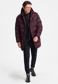 Giubbotto piumino bordeaux con cappuccio, design trapuntato, chiusura con zip e tasche laterali. Abbinato a jeans scuri e stivali neri alla caviglia.