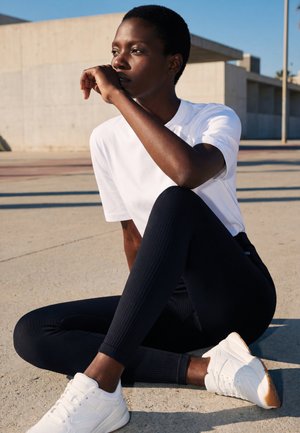 Osoba siedząca, z krótkimi włosami, ubrana w białą koszulkę, czarne legginsy i białe trampki, odpoczywająca na zewnątrz na betonowej nawierzchni.