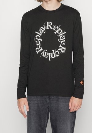 Osoba nosící černé tričko s dlouhým rukávem, na hrudi s opakujícím se textem „Replay“ ve tvaru kruhu a malým oranžovým logem Replay na rukávu.