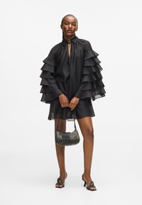 Μαύρο φόρεμα με στρώσεις βολάν στα μανίκια, κολαριστό ντεκολτέ και μήκος μέχρι το γόνατο. Το μοντέλο κρατάει μια φλυαρία τσάντα και φοράει πέδιλα.