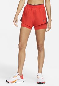 Persona vestida con pantalones cortos atléticos rojos en capas y zapatillas blancas de running con acentos en naranja y rosa, de pie contra un fondo blanco.