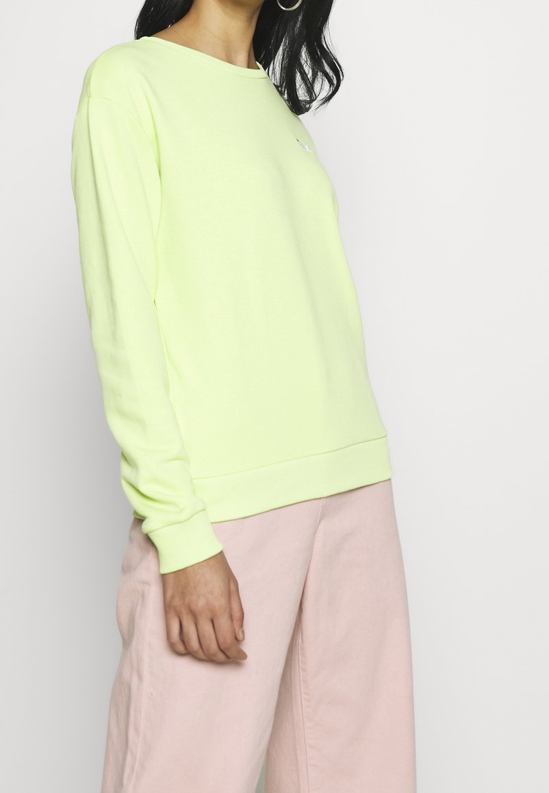 Felpa verde neon con scollo rotondo, polsini e orlo a costine. Abbinata a pantaloni rosa chiaro a gamba larga, che mostrano una texture liscia e morbida.