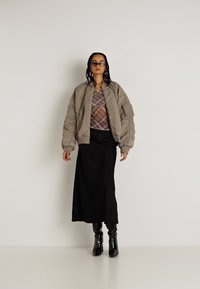 Jaqueta bomber acolchoada cinza claro, blusa transparente com padrão, saia maxi preta texturizada e botas pretas até ao joelho. O modelo está de pé contra um fundo branco.