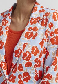 Blazer floral en tissu bleu clair avec motifs floraux rouges et beiges. Superposé à un haut orange texturé et côtelé.