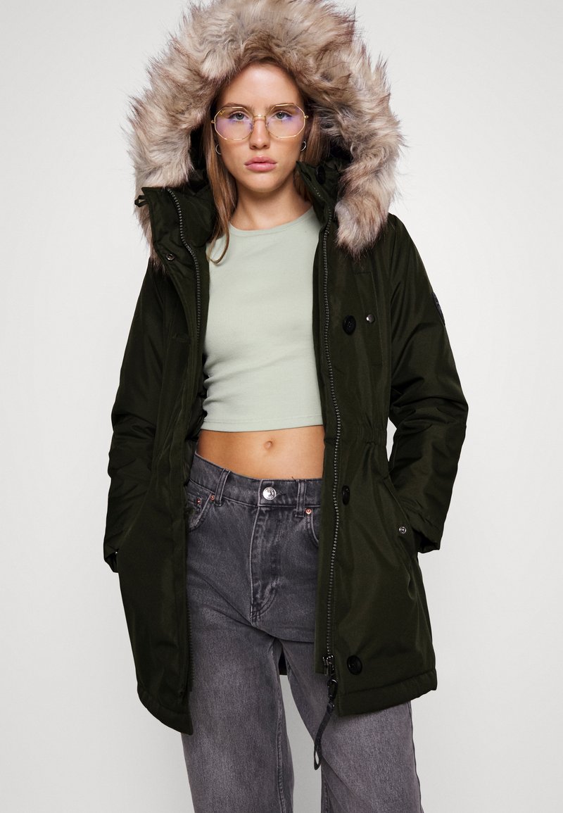 Donna che indossa occhiali, parka verde scuro con cappuccio foderato di pelliccia, top corto verde chiaro e jeans grigi a vita alta, in piedi con le mani nelle tasche.