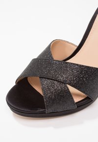 Sandale noire à bout ouvert avec des brides croisées scintillantes et une doublure intérieure beige sur fond blanc.