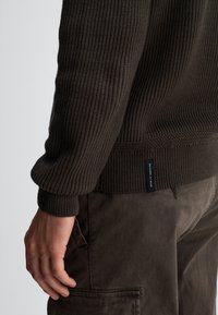 Dunkelbrauner, strukturierter Strickpullover mit gerippten Bündchen und Saum, versehen mit einem kleinen Etikett an der Seite. Kombiniert mit dunklen Hosen.