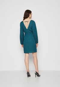 Robe turquoise avec de longues manches bouffantes, un dos en V profond, un tissu texturé et une ceinture à la taille. Portée avec des chaussures à talons hauts noires.