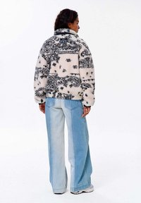 Žena otočená chrbtom, oblečená v čierno-bielej vzorovanej fleecovej bunde, svetlomodrých širokých nohaviciach a bielych teniskách na bielom pozadí.