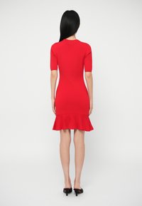 Γυναίκα με μακριά μαύρα μαλλιά που φοράει ένα κολλητό κόκκινο φόρεμα με κοντά μανίκια και φουσκωτό τελείωμα, στέκεται με την πλάτη στραμμένη προς το φακό σε λευκό φόντο.