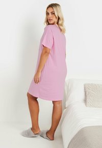 Rosa bomullsnattklänning med korta ärmar, knälång, avslappnad passform. Modell som bär fluffiga grå tofflor, står bredvid en prydligt bäddad säng.