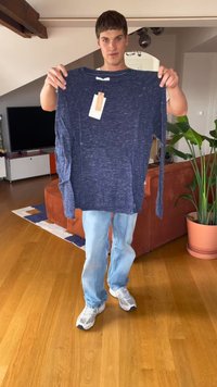 Młody mężczyzna stojący w salonie trzyma niebieską koszulę z długim rękawem z metkami, ubrany w jasne dżinsy i białe sneakersy.