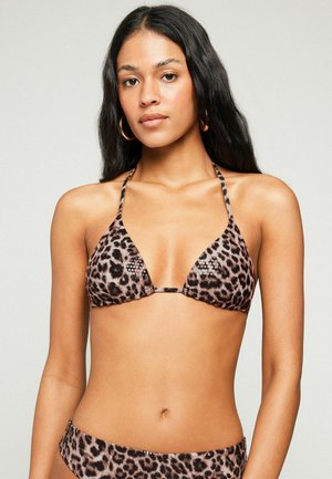 Junge Frau mit langen dunklen Haaren, die goldene Creolen und einen Leoparden-Bikini trägt, blickt nach vorne vor einem einfarbigen Hintergrund.