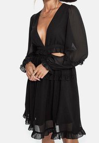 Kvinna i en svart långärmad klänning med djup V-ringning, volangdetaljer vid midjan, ärmarna och fållen, prydd med guldringar och halsband.