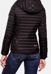 Žena v těsně padnoucí černé prošívané bundě s kapucí a modrých džínách, zobrazená zezadu na jednobarevném pozadí.