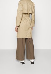 Beige trenčkot s přiléhavým pasem a širokými klopami, s podelním klopovým detail na zádech. Spárováno s hnědými kalhotami s širokými nohavicemi a bílými botami na blokovém podpatku.