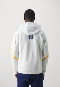 Witte hoodie met marineblauwe strepen op de achterkant en gele accenten op de mouwen. Heeft een ontspannen pasvorm en ribgebreide manchetten.