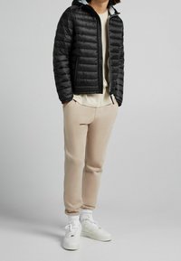 Černá prošívaná bunda s kapucí, prošívaný design a přední zip; béžové tepláky; bílé tenisky s texturovanou podrážkou.