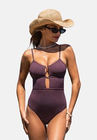 Maillot de bain une pièce bordeaux avec un motif texturé, présentant un design découpé et des bretelles fines. Accessoirisé avec un chapeau en paille et des lunettes de soleil.