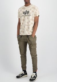 Camiseta beige con patrón de camuflaje y logo negro, combinada con pantalones cargo verde olivo con cremalleras y bolsillos. Zapatillas negras y blancas.