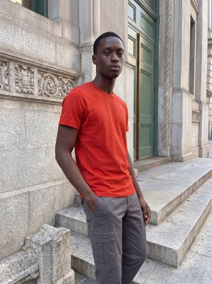 Ung mand i rød t-shirt og grå cargobukser står med hænderne i lommerne ved udsmykkede stentrapper og grønne døre.