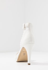 Bílá lodička na vysokém podpatku s hladkým koženým povrchem, přezkou a vertikálním zdobením na zadní straně. Velikost označena jako 39 na podrážce.