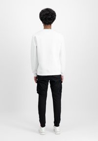 Sweat-shirt blanc avec poignets et ourlet côtelés, assorti à un pantalon cargo noir à poches, complété par des baskets blanches. Fond uni.