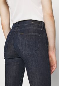 Vaqueros de mezclilla azul oscuro con corte slim, que presentan un logo en el bolsillo trasero, costuras sutiles y diseño estándar de cinco bolsillos.