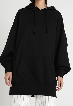 Frau trägt einen übergroßen schwarzen Hoodie mit Fronttasche und Kordeln, kombiniert mit weißen Hosen mit dünnen schwarzen vertikalen Streifen.