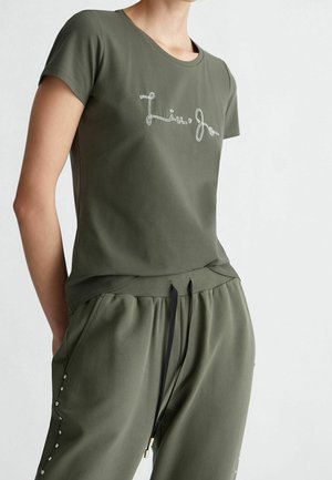 Frau trägt ein olivgrünes Kurzarmhemd mit "Liu Jo" Stickerei und passende Hose mit seitlichen Knopfdetails und schwarzem Kordelzug.
