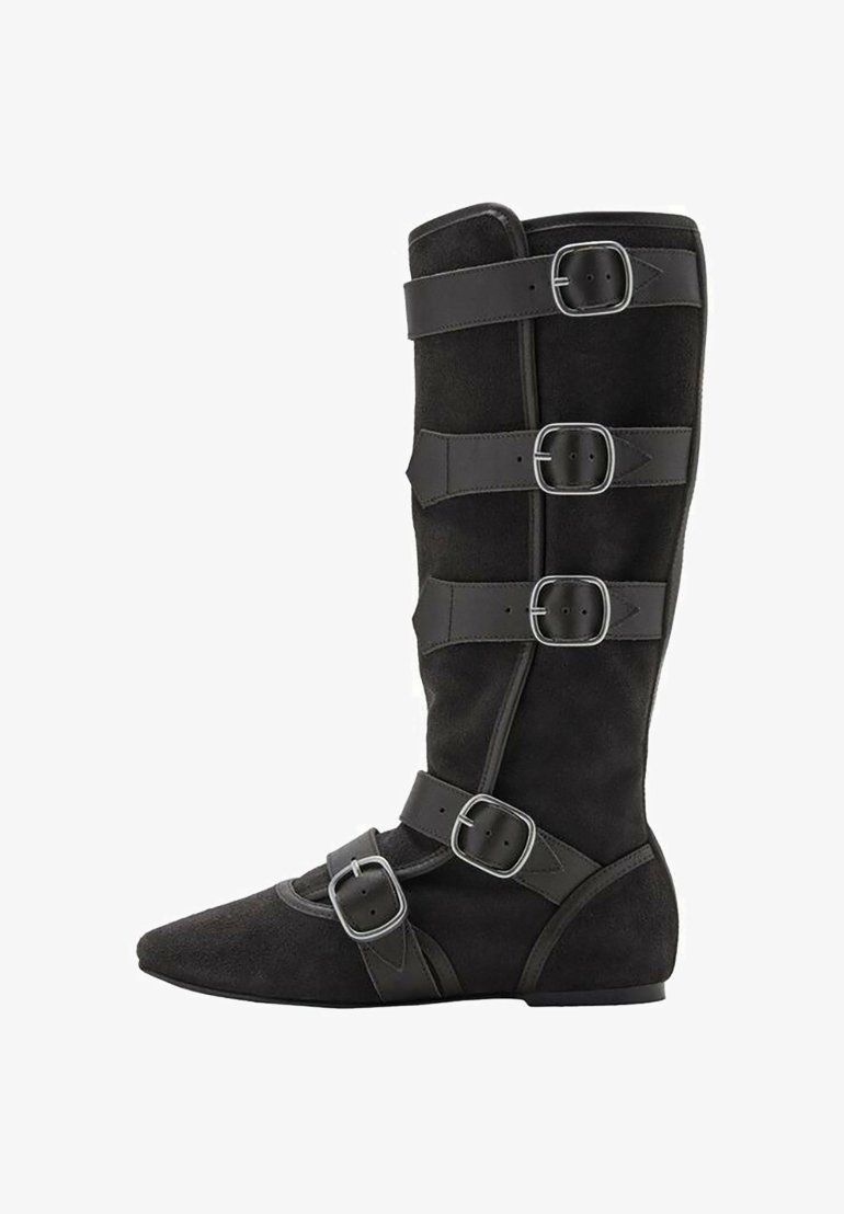 Bota de ante negra hasta la rodilla con punta afilada, que presenta múltiples correas y detalles de hebilla a lo largo del eje y el tobillo. Diseño de suela plana.