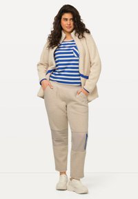 Beige Fleecejacke mit blauen Akzenten, getragen über einem blau-weiß gestreiften Hemd und beigen Hosen mit hervorgehobenen Taschen und Texturen.